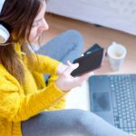 Młoda Nowożytna kobieta siedzi na podłoga w sypialni i używa telefon robić zakupy online przy użyciu karty kredytowej